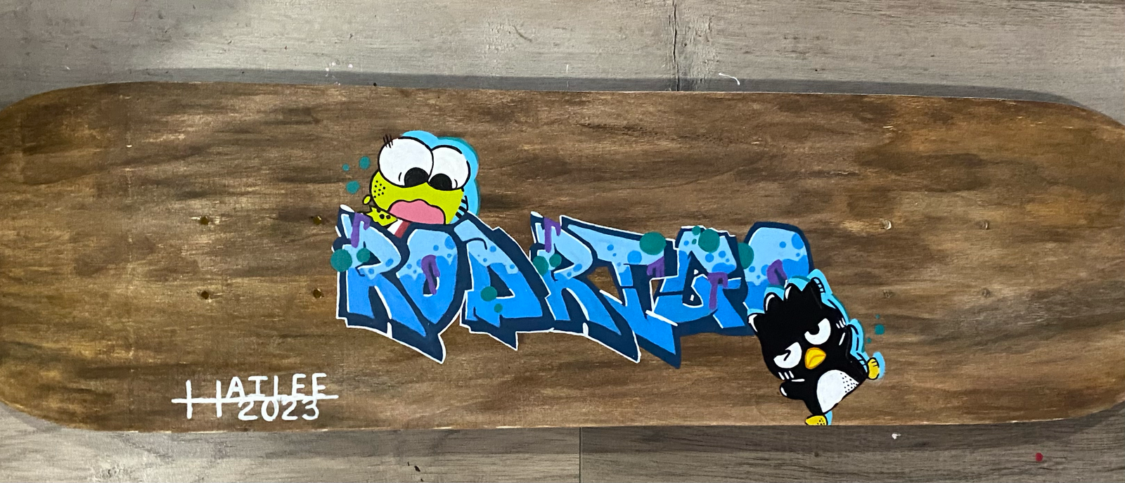  stained wood skateboard with the name Rodrigo written on it. on top of the letters R and O is the character Keroppi from sanrio. on the right side close to the last letter in the name is the sanrio character Badtz Maru.